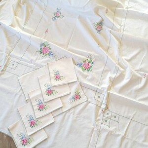 May include: A white tablecloth with embroidered floral designs in shades of pink, green, and blue. The tablecloth is accompanied by eight matching napkins with the same floral design.