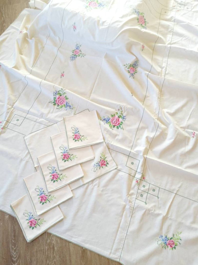 May include: A white tablecloth with embroidered floral designs in shades of pink, green, and blue. The tablecloth is accompanied by eight matching napkins with the same floral design.