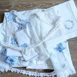 Könnte beinhalten: Eine Sammlung weißer und blauer Vintage-Bettwäsche, darunter Kissenbezüge, Deckchen und Spitzenbesatz. Einige Teile weisen blaue Blumenstickereien und Schmetterlingsmuster auf. Die Artikel sind auf einer Holzoberfläche angeordnet.