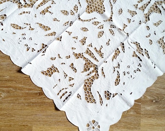 May include: White, intricately cut lace tablecloth with a scalloped edge. The design features floral and geometric patterns, creating a delicate and elegant aesthetic. The tablecloth is placed on a wooden surface.