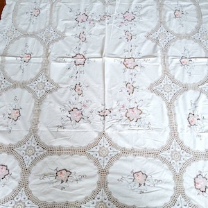 Op de afbeelding: Witte tafelkleed met delicate gehaakte kant en geborduurde bloemenmotieven in roze en bruine tinten.