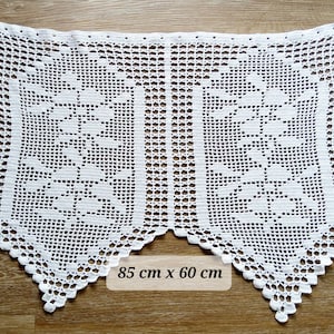 Peut inclure: Un napperon rectangulaire en crochet blanc présentant un motif floral symétrique avec des sections en maille ajourée. La pièce mesure 85 cm sur 60 cm comme indiqué par le texte en surimpression. Les bords ont une finition festonnée.
