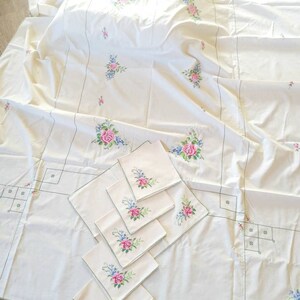 May include: A white tablecloth with embroidered floral designs in pink, blue, and green. The tablecloth is accompanied by six matching napkins with the same floral design.