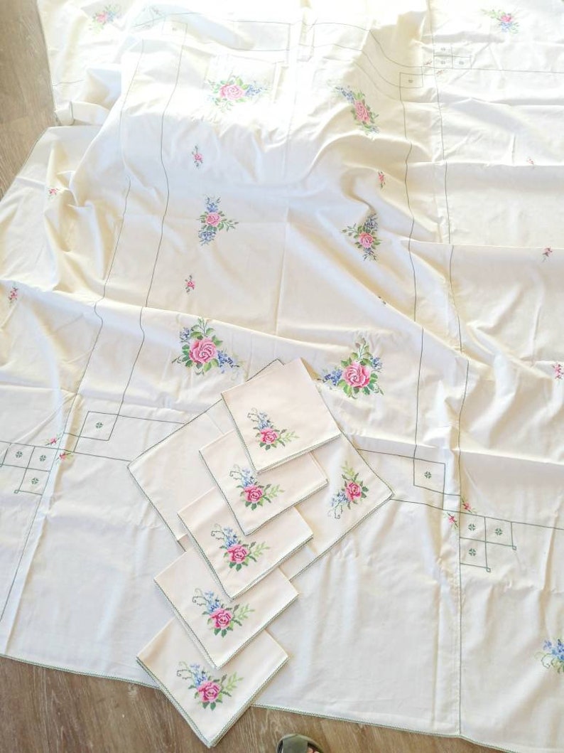 May include: A white tablecloth with embroidered floral designs in pink, blue, and green. The tablecloth is accompanied by six matching napkins with the same floral design.