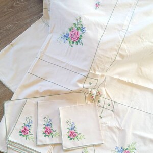 May include: A set of white embroidered table linens with a floral design. The set includes a tablecloth, placemats, and napkins. The floral design features pink and blue flowers with green leaves.