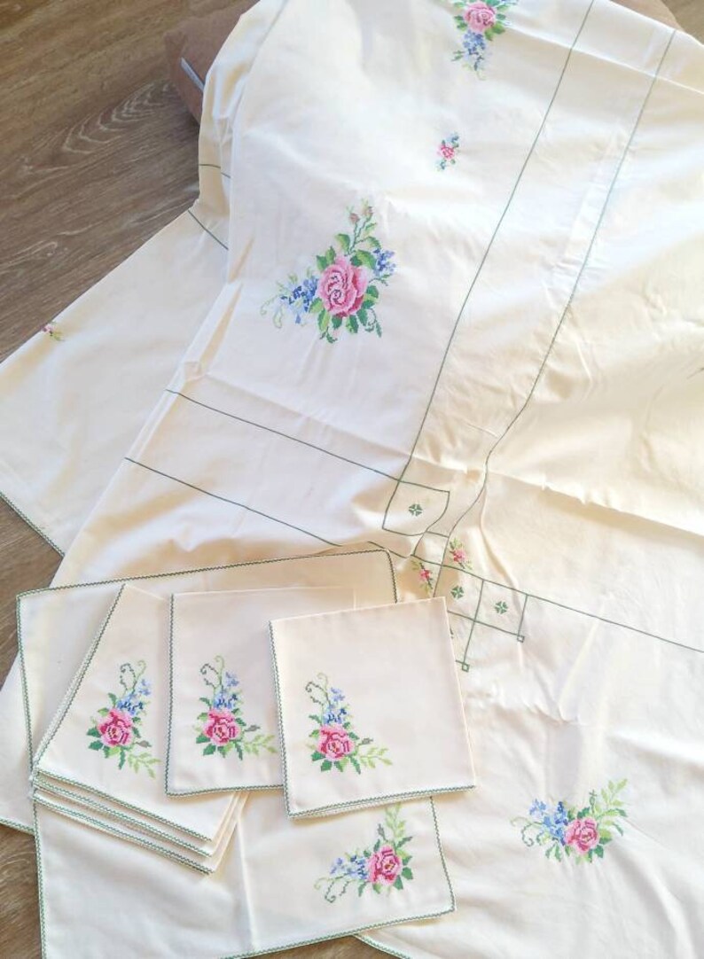 May include: A set of white embroidered table linens with a floral design. The set includes a tablecloth, placemats, and napkins. The floral design features pink and blue flowers with green leaves.