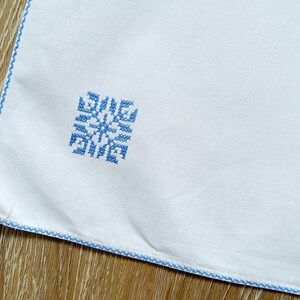 Puede incluir: Tela blanca con un dise&ntilde;o de copo de nieve bordado en azul en la esquina. Los bordes est&aacute;n ribeteados con un borde cosido azul. La tela est&aacute; sobre una superficie de madera.