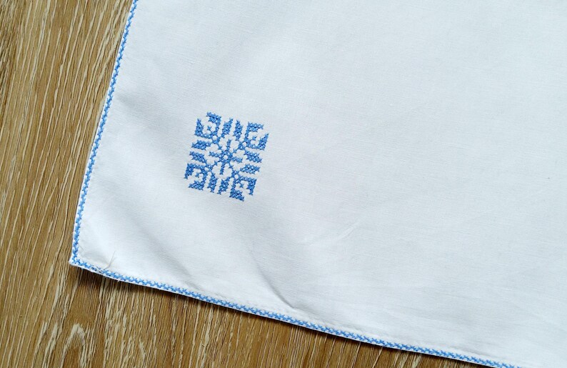 Puede incluir: Tela blanca con un dise&ntilde;o de copo de nieve bordado en azul en la esquina. Los bordes est&aacute;n ribeteados con un borde cosido azul. La tela est&aacute; sobre una superficie de madera.