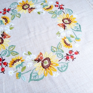 Könnte beinhalten: Eine weiße Leinen-Tischdecke mit einem gestickten Sonnenblumenkranz-Design. Der Kranz zeigt gelbe Sonnenblumen, grüne Blätter, rote Beeren und weiße Blüten.