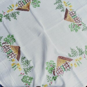 May include: White linen tablecloth with embroidered floral and house designs in shades of green, brown, yellow, and red.