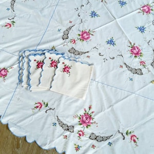 May include: A white tablecloth with a scalloped edge and embroidered floral designs in pink, blue, and green. The tablecloth is accompanied by four matching napkins with scalloped edges and embroidered floral designs.
