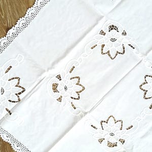 Könnte beinhalten: Weißes Leinentischtuch mit ausgeschnittenen floralen Designs und zarter Spitzenborte. Das Tischtuch weist ein sich wiederholendes Muster stilisierter Blumen auf und verleiht jeder Tafel einen Hauch von Eleganz.