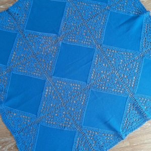 Puede incluir: Un edredón cuadrado azul hecho a mano con un diseño de patchwork. El edredón presenta cuadrados azules lisos intercalados con cuadrados de encaje calado. El edredón se muestra sobre una superficie de madera.