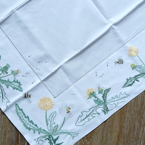 Könnte beinhalten: Weißes Tischtuch mit gestickten Details. Der Rand zeigt grüne Löwenzahnblätter, gelbe und weiße Löwenzahnblüten und kleine Bienen. Das Tischtuch ist gefaltet, mit einer gepunkteten Linienbordüre.