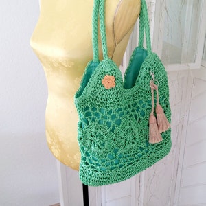 Puede incluir: Un bolso de mano de crochet verde con un botón en forma de flor y borlas. El bolso está colgado de un maniquí amarillo.
