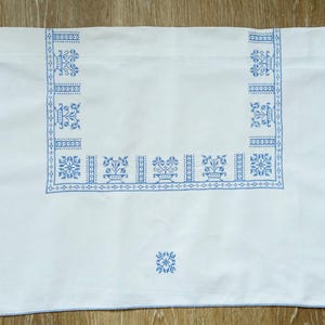 Puede incluir: Mantel rectangular blanco con patrones florales y geom&eacute;tricos bordados en azul. El borde presenta un dise&ntilde;o repetitivo de jarrones y flores estilizadas. Peque&ntilde;os motivos florales en cada esquina.