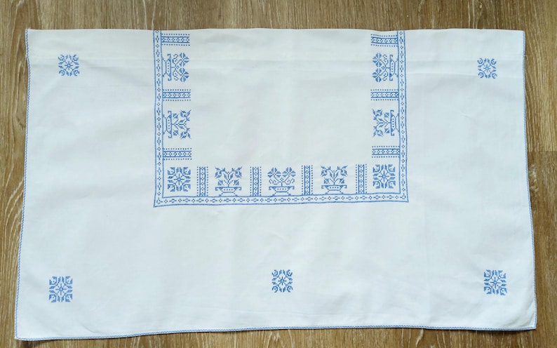 Puede incluir: Mantel rectangular blanco con patrones florales y geom&eacute;tricos bordados en azul. El borde presenta un dise&ntilde;o repetitivo de jarrones y flores estilizadas. Peque&ntilde;os motivos florales en cada esquina.