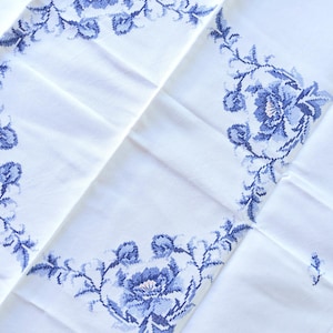 Könnte beinhalten: Weißes Tischtuch mit blauem Blumenmuster. Die Stickerei zeigt detaillierte blaue Blumen und Blätter, die einen klassischen und eleganten Look erzeugen. Das Tuch ist gefaltet und zeigt das Muster in Abschnitten.