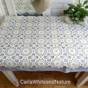 Könnte beinhalten: Eine cremefarbene, gehäkelte Spitzendecke mit floralem Muster bedeckt einen Tisch. Die Tischdecke ist über einem hellblauen Stoff platziert. Der Tisch steht vor einer weißen Backsteinmauer. Der Text "CarlaWhiteandNature" ist sichtbar.
