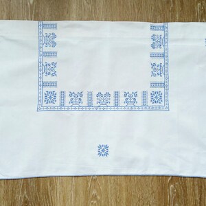 Puede incluir: Mantel rectangular blanco con bordados azules de motivos florales y geom&eacute;tricos. El borde y el dise&ntilde;o central presentan motivos detallados de estilo punto de cruz. El mantel est&aacute; colocado sobre una superficie de madera.