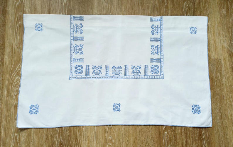 Puede incluir: Mantel rectangular blanco con bordados azules de motivos florales y geom&eacute;tricos. El borde y el dise&ntilde;o central presentan motivos detallados de estilo punto de cruz. El mantel est&aacute; colocado sobre una superficie de madera.