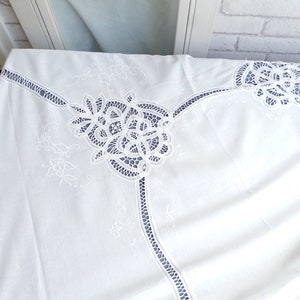 Puede incluir: Mantel de mesa de algod&oacute;n blanco con delicados detalles de encaje bordado. El encaje presenta intrincados patrones florales y un borde festoneado.