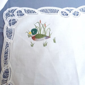 Puede incluir: Mantel de encaje blanco con un dise&ntilde;o de pato bordado en verde, marr&oacute;n y amarillo.