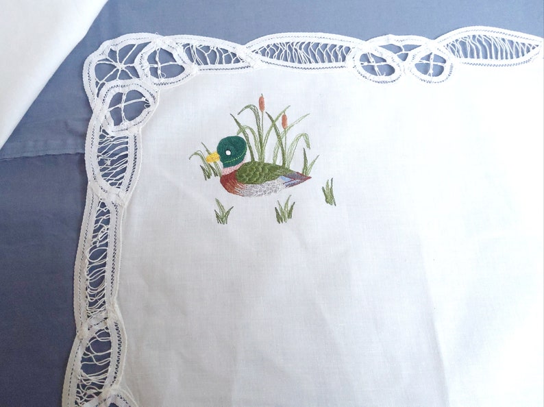 Puede incluir: Mantel de encaje blanco con un dise&ntilde;o de pato bordado en verde, marr&oacute;n y amarillo.