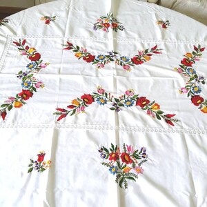 Könnte beinhalten: Runder weißer Tischdecken mit gestickten Blumendesigns in Rot, Gelb, Blau und Grün. Die Tischdecke hat einen zarten Spitzenrand.