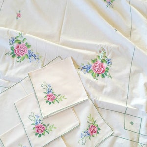 May include: Set of four white linen napkins with green embroidered floral designs. The napkins are folded and arranged on a white tablecloth with a similar floral design.