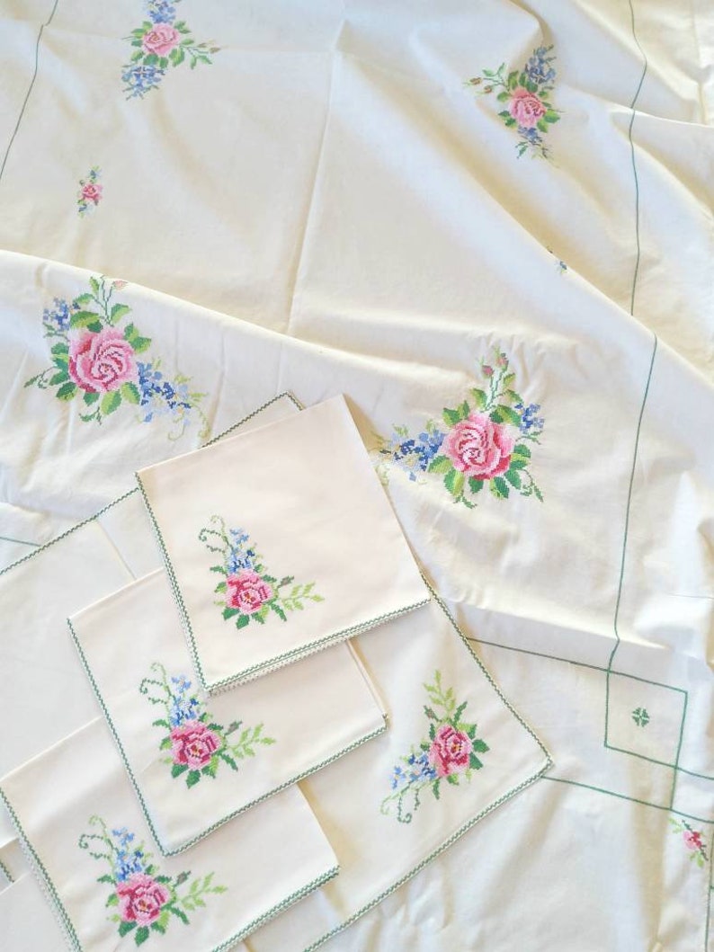 May include: Set of four white linen napkins with green embroidered floral designs. The napkins are folded and arranged on a white tablecloth with a similar floral design.