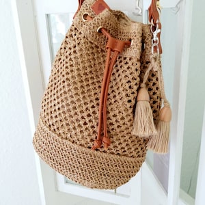 May include: A brown crocheted bucket bag with brown leather straps and tassels. The bag has a drawstring closure and a small heart-shaped tag.