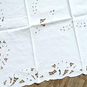 May include: A white, rectangular tablecloth with detailed floral cutwork around the borders. The design includes intricate flower and leaf patterns, giving a delicate and elegant appearance. The tablecloth is made of a soft, white fabric.