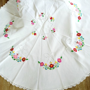 Op de afbeelding: Witte tafelkleed met een kantversiering en geborduurde bloemenmotieven in tinten roze, rood, blauw, geel en groen.