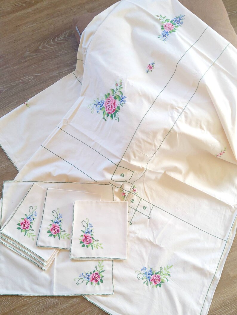 May include: A white tablecloth with green embroidered trim and floral designs. The tablecloth is accompanied by six matching napkins with the same floral design.