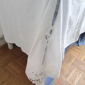 Puede incluir: Mantel blanco con delicados dise&ntilde;os florales bordados y ribete de encaje. El mantel est&aacute; drapeado sobre una superficie de madera.