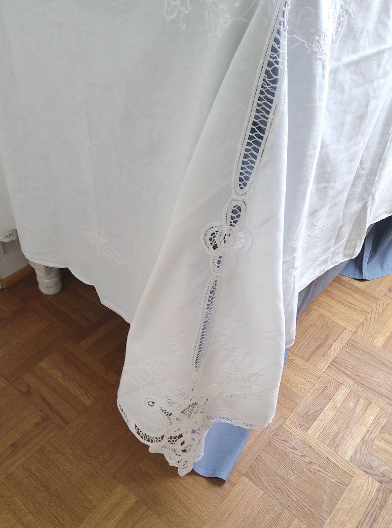 Puede incluir: Mantel blanco con delicados dise&ntilde;os florales bordados y ribete de encaje. El mantel est&aacute; drapeado sobre una superficie de madera.