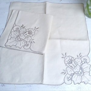 以下が含まれることがあります： 白いリネン製のナプキン2枚。角に刺繍された花柄が施されています。ナプキンは折り畳まれ、白い表面に置かれています。