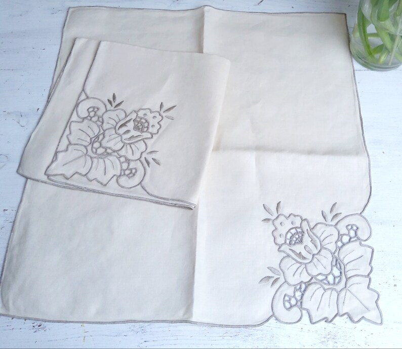 以下が含まれることがあります： 白いリネン製のナプキン2枚。角に刺繍された花柄が施されています。ナプキンは折り畳まれ、白い表面に置かれています。
