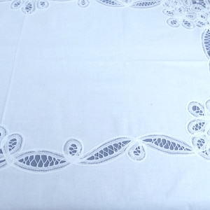 Puede incluir: Mantel blanco con un delicado patr&oacute;n de encaje alrededor de los bordes. El encaje presenta intrincados dise&ntilde;os florales y remolinos.
