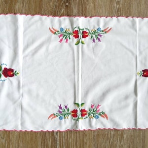 以下が含まれることがあります： 赤、ピンク、黄色、青、緑の刺繍された花柄が施された、白地に波状の縁取りのあるテーブルランナーです。