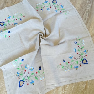 Puede incluir: Un mantel de mesa de lino beige con diseños florales bordados en azul, verde, rojo y amarillo. Las flores son estilizadas y tienen un centro en forma de corazón.