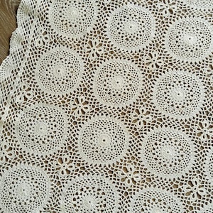 Peut inclure: Une nappe en crochet blanc cassé présentant un motif répétitif de grands cercles reliés par de petites fleurs à quatre pétales. Le tissu possède un bord festonné et un design ajouré complexe sur l'ensemble de la pièce textile.