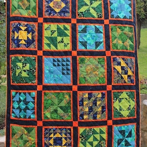 Peut inclure: Une courtepointe avec un fond noir et des bordures orange. La courtepointe est composée de 16 carrés, chacun avec un motif différent de tissu vert, bleu et jaune. Les motifs sont géométriques et abstraits.