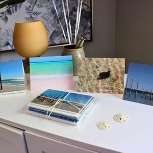May include: A collection of coastal-themed greeting cards featuring beach and ocean scenes. The cards are stacked and tied with twine, with individual cards displaying images of piers, water, and sand. Two sand dollars are also present.