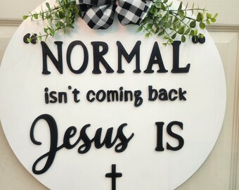 May include: A round, white wooden sign with the text "NORMAL isn't coming back Jesus IS" in black lettering. The sign is decorated with a black and white checkered bow, greenery, and a small cross.