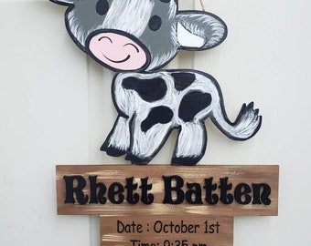Puede incluir: Un letrero de madera con una vaca de dibujos animados pintada y el texto "Rhett Batter" en la parte superior. El letrero también incluye la fecha de nacimiento, la hora, el peso y la longitud del bebé: 1 de octubre, 9:35 pm, 6 libras 5 onzas, 19 1/4 pulgadas.