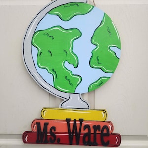 May include: A wooden globe-shaped sign with a light blue background and green continents. The globe sits atop a stack of three books in yellow, orange, and red. The name "Ms. Ware" is displayed in black letters.