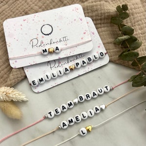 Könnte beinhalten: Drei Armbänder mit weißen Perlen und schwarzen Buchstaben. Die Armbänder buchstabieren "TEAM BRAUT", "AMELIA" und "EMILIA PABLO". Jedes Armband hat einen goldenen Herzanhänger.