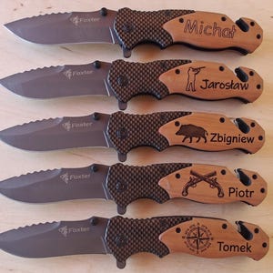 Puede incluir: Cinco navajas plegables con hojas de metal gris marcadas Foxter, mangos negros texturizados y empuñaduras de madera clara. Cada empuñadura presenta diseños grabados con láser únicos que incluyen nombres como Michał, Jarosław, Zbigniew, Piotr y Tomek.